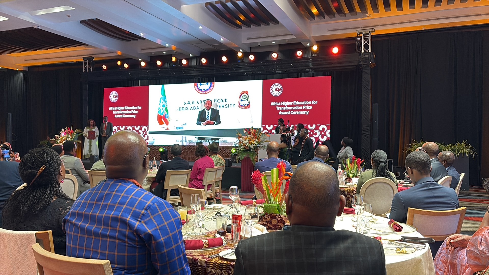 A view of the event space from Heather's perspective, sat at a roundtable, looking at the stage which has a large screen as its backdrop, displaying the name of the event.