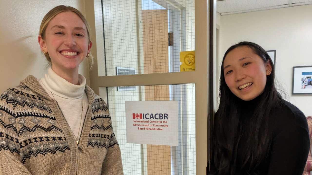 Annelise Jahnke (left) with Megan Toi (right), photographed in the ICACBR offices, standing either side of a window with the ICACBR logo printed on it.