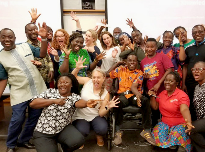 A diverse group of smiling adults pose together in a lively, celebratory group photo. Some people are raising their hands, pointing, or making playful gestures, creating a joyful and inclusive atmosphere. One individual at the front uses a wheelchair. 