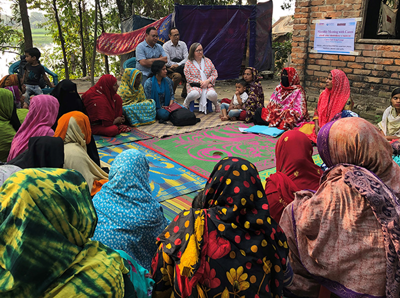 A community meeting held outdoors in a rural village setting. Women in colourful saris sit in a circle while facilitators engage in discussion, with a banner posted on a nearby brick wall.