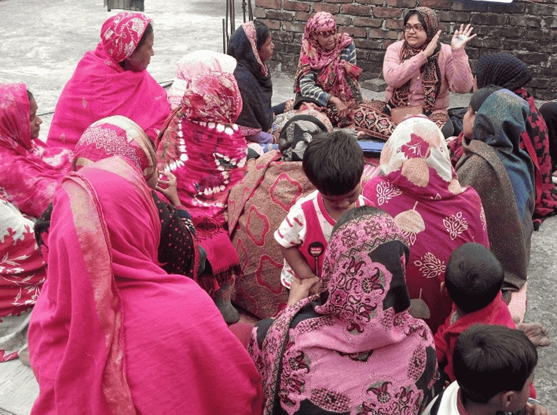 Women and children are gathered and sitting listening to a woman speaker