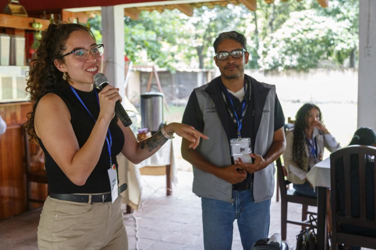 Valeria stands, holding a microphone, smiling and addressing the group.