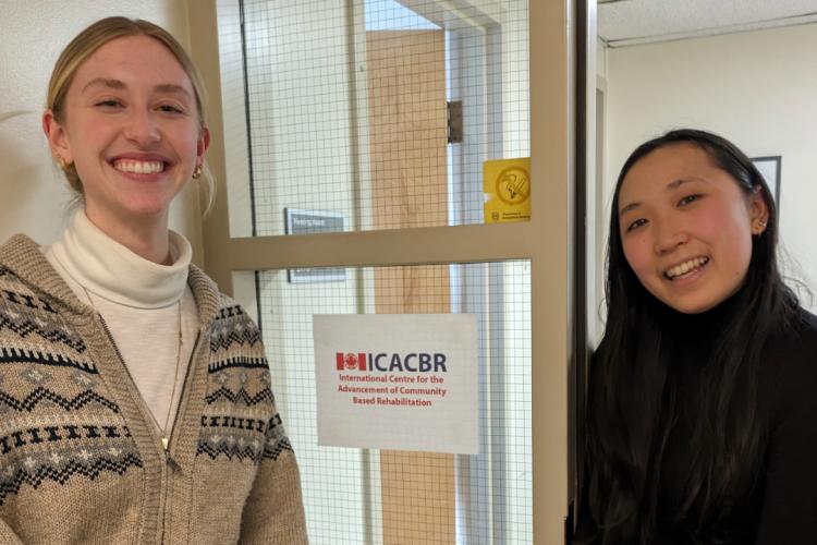 Annelise Jahnke (left) with Megan Toi (right), photographed in the ICACBR offices, standing either side of a window with the ICACBR logo printed on it.