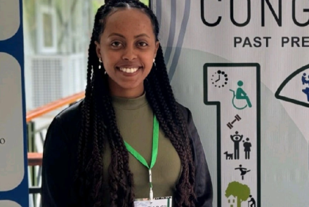 Rahel Olana stands, wearing a black blazer on top of a green tee. She is smiling at the camera.