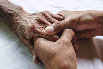 The hand of an elderly person is held with two hands of a younger person.